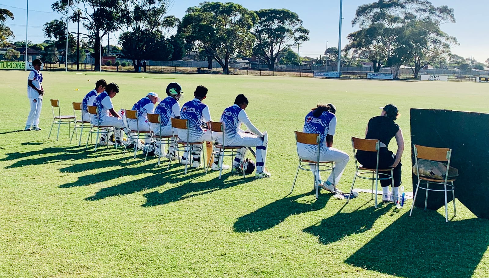 A VECL team waiting to bat: players seated in a row of chairs at the boundary in afternoon sun, pads on, watching the match.